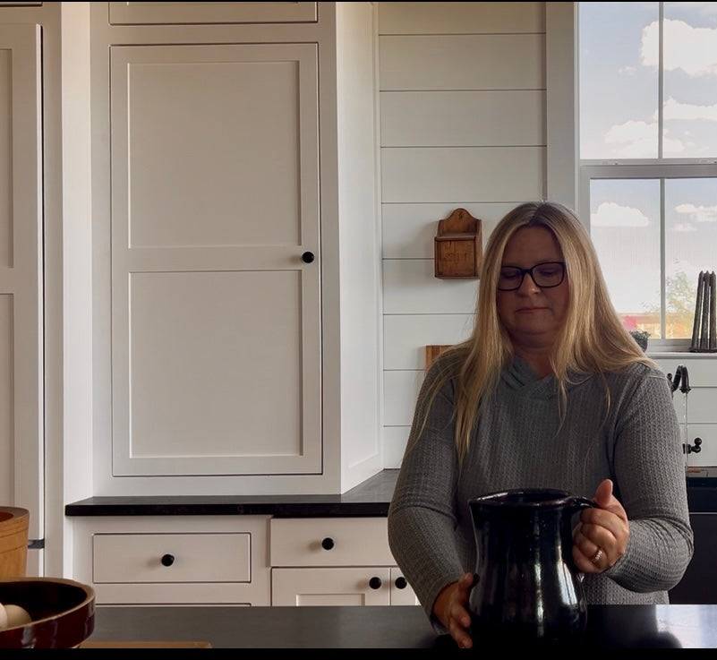 Woman holding a black pot in a kitchen with white cabinets and a window in the background.